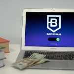 Laptop exibindo uma interface de conexão blockchain sobre uma mesa, ao lado de livros e cédulas de dólar.