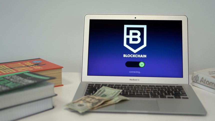 Laptop exibindo uma interface de conexão blockchain sobre uma mesa, ao lado de livros e cédulas de dólar.