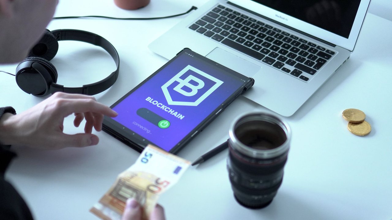 Tablet exibindo uma transação de moeda digital ao lado de um laptop e cédulas de euro.