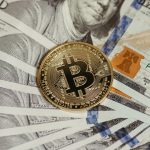 Moeda de Bitcoin apoiada sobre uma pilha de cédulas de cem dólares.