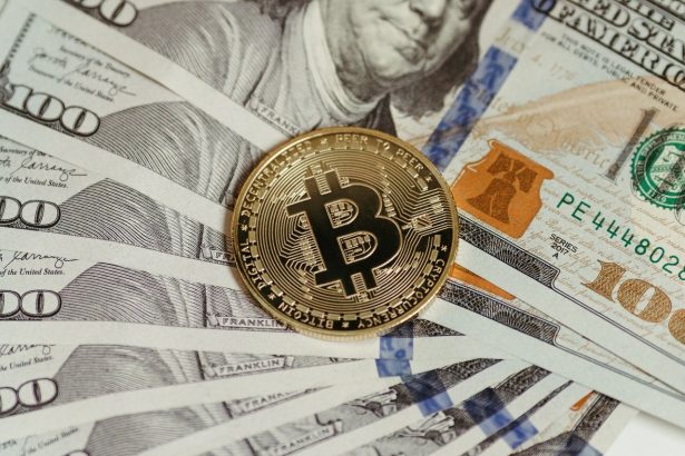 Moeda de Bitcoin apoiada sobre uma pilha de cédulas de cem dólares.