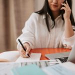 Mulher sentada à mesa, fazendo anotações enquanto fala ao telefone e trabalha em um laptop.