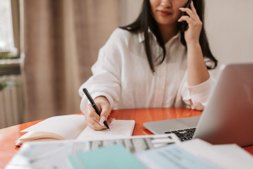 Mulher sentada à mesa, fazendo anotações enquanto fala ao telefone e trabalha em um laptop.