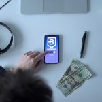 Celular exibindo um aplicativo de blockchain ao lado de cédulas, fones de ouvido e um caderno.