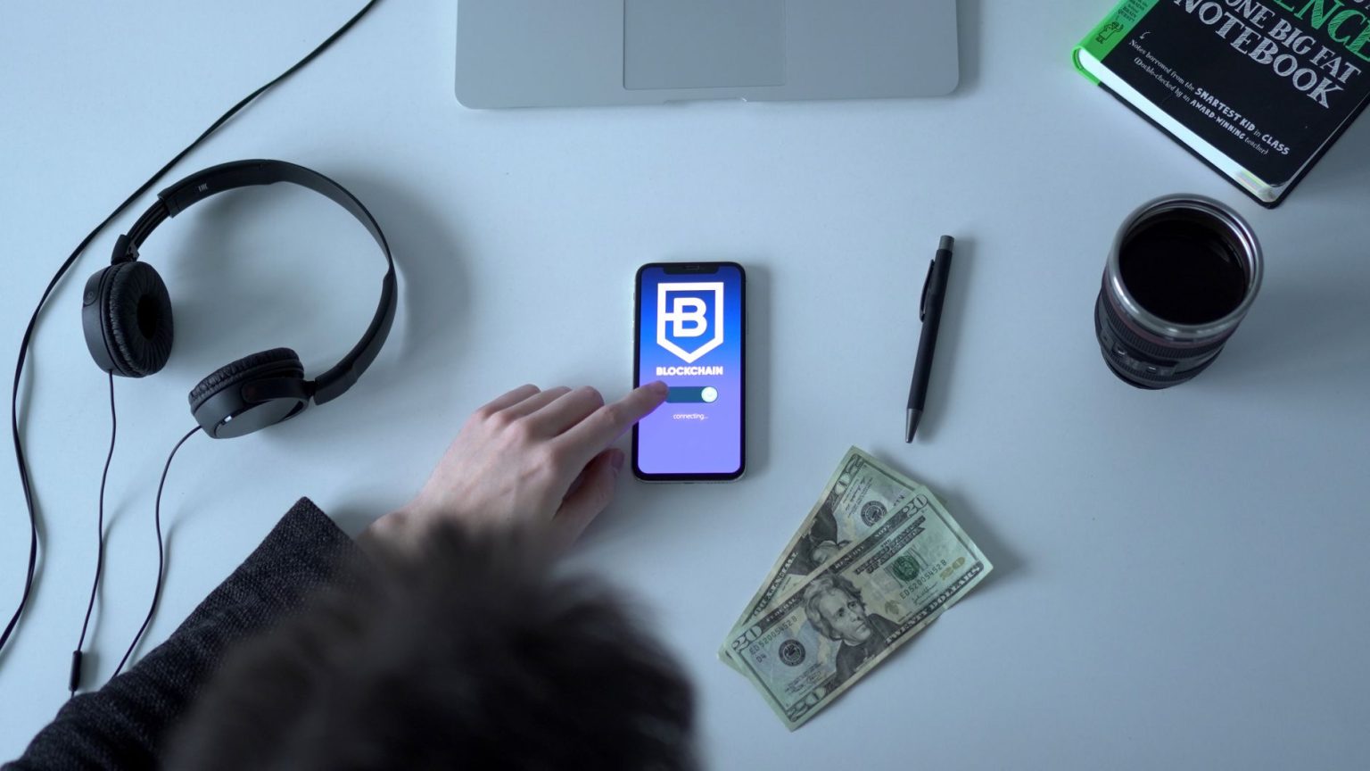 Celular exibindo um aplicativo de blockchain ao lado de cédulas, fones de ouvido e um caderno.