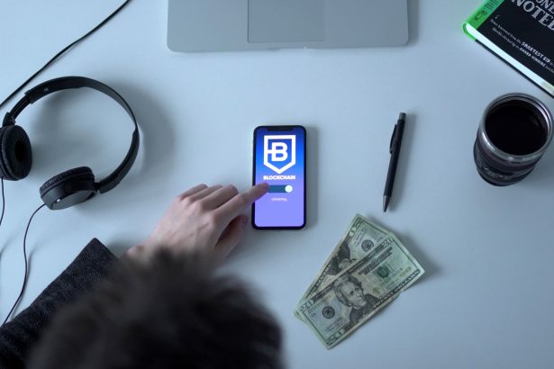 Celular exibindo um aplicativo de blockchain ao lado de cédulas, fones de ouvido e um caderno.
