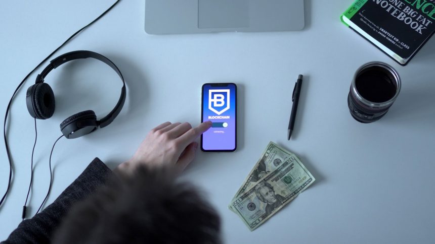 Celular exibindo um aplicativo de blockchain ao lado de cédulas, fones de ouvido e um caderno.